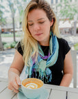 Vibrant blue + greens hand-painted scarf wrapped on a model. The model is enjoying a cup of coffee.