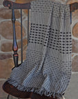 The women's handwoven gray shawl draped over a chain with a stone wall behind it. The eyelash fringe is also in view.