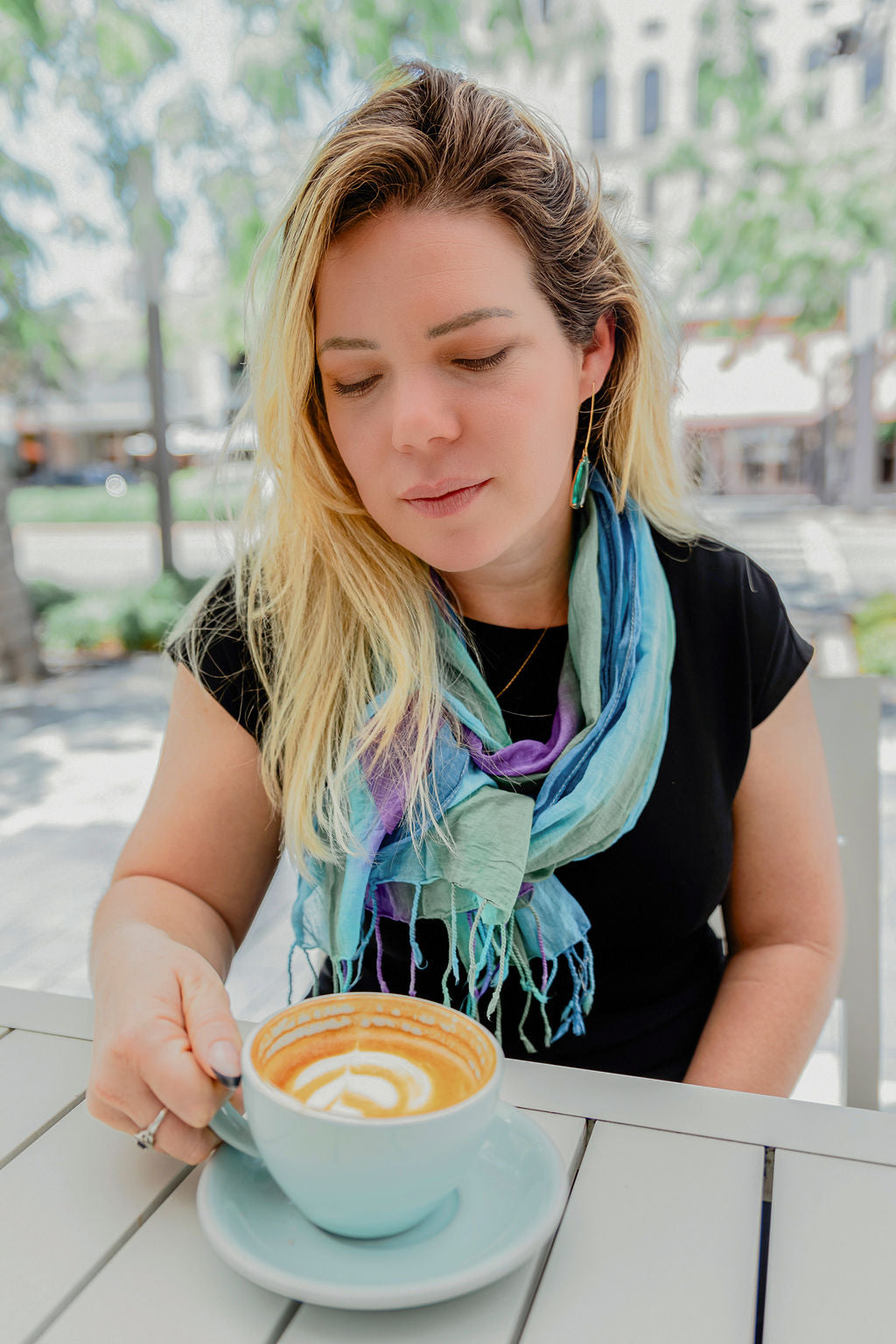 Vibrant blue + greens hand-painted scarf wrapped on a model. The model is enjoying a cup of coffee.