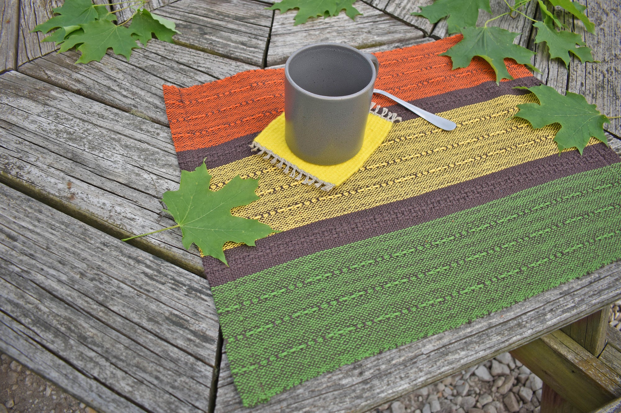 Handwoven fall placemat sitting on a picnic table. There are green leaves and mug in view.