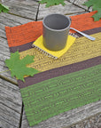 Handwoven fall placemat sitting on a picnic table. There are green leaves and mug in view.