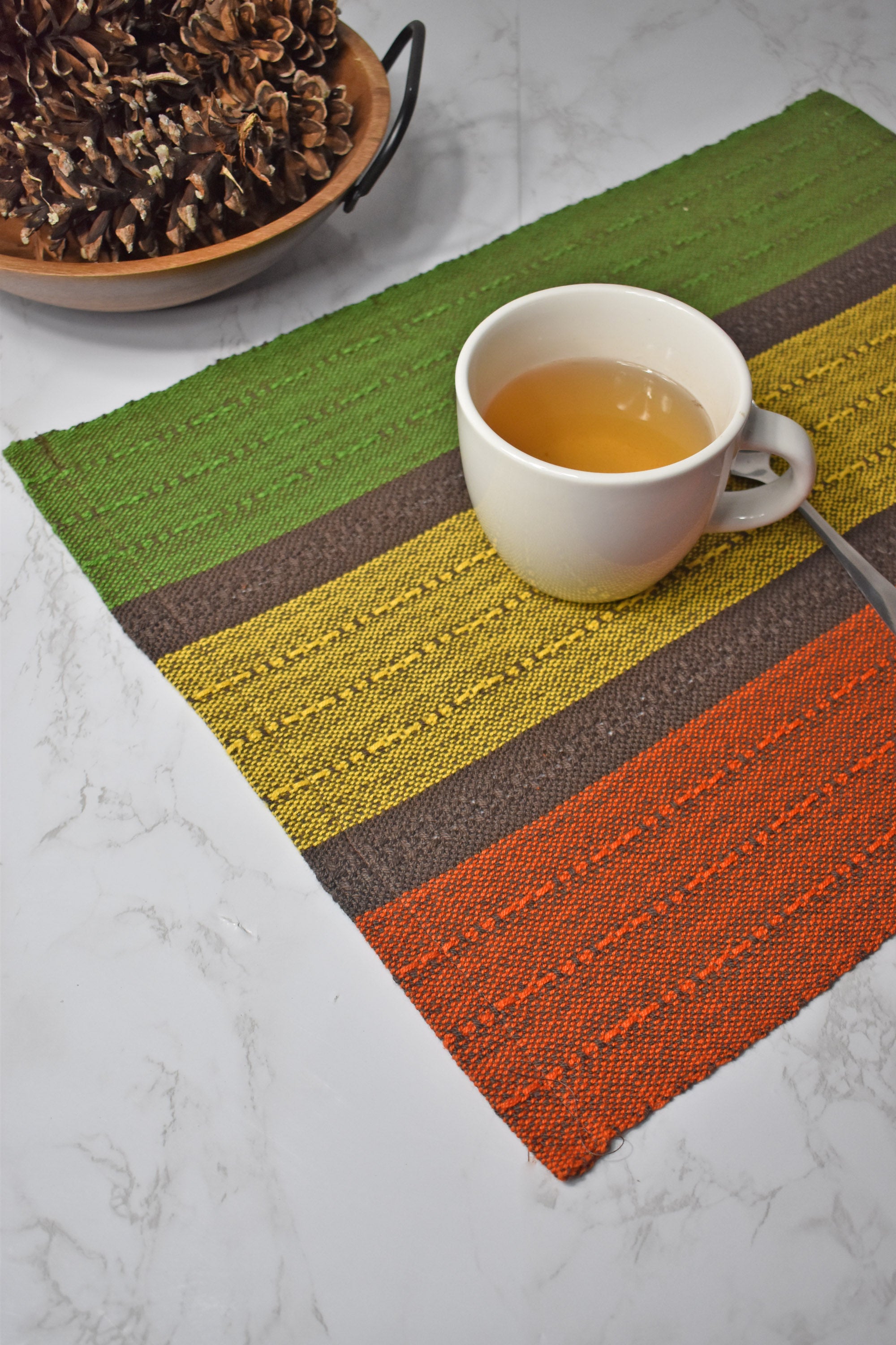 Fall inspired placemat on a marble table with a cup of tea on top of it. Bowl of pine cones on the top left of photo.