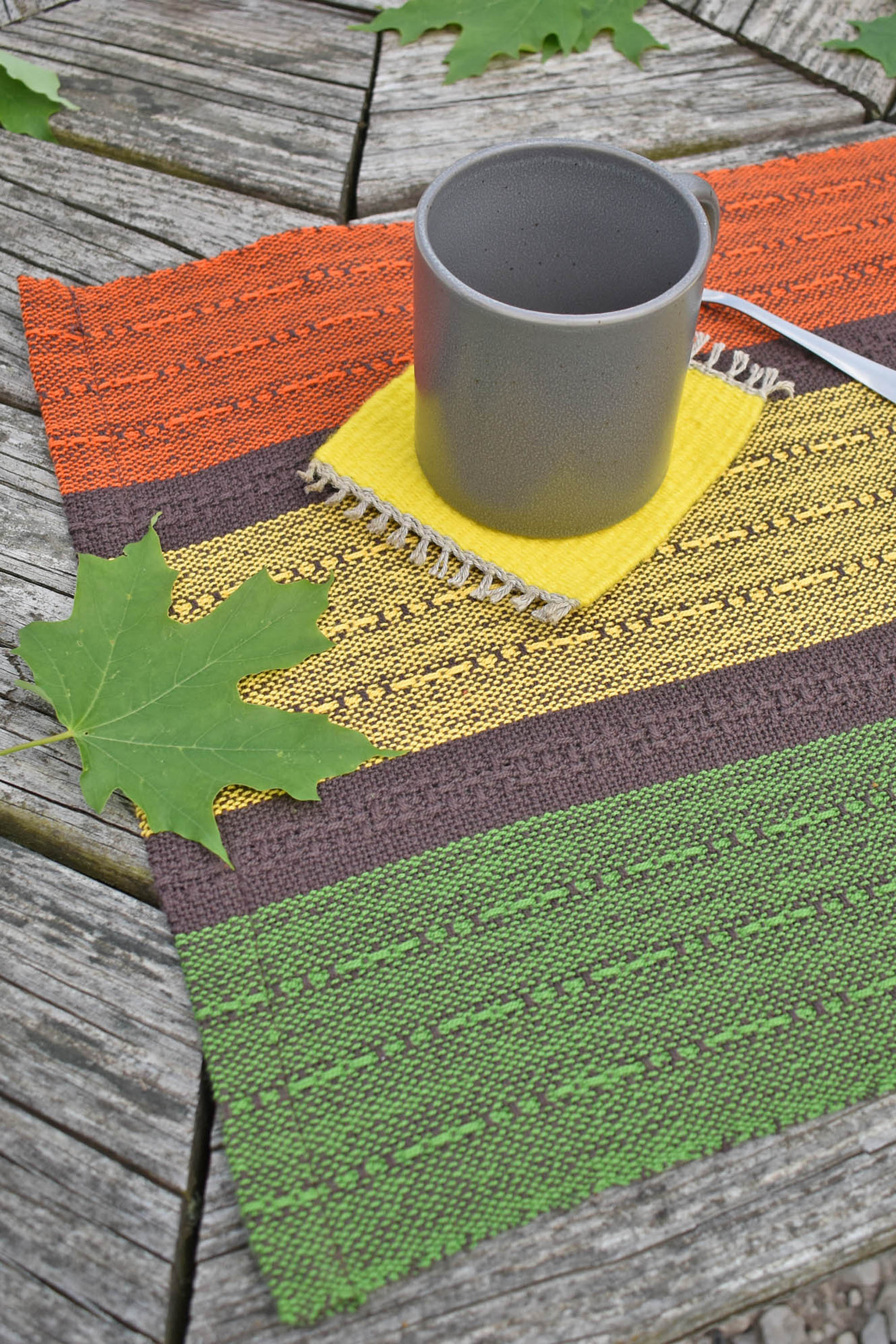 Handwoven fall placemat sitting on a picnic table. There are green leaves and mug in view.