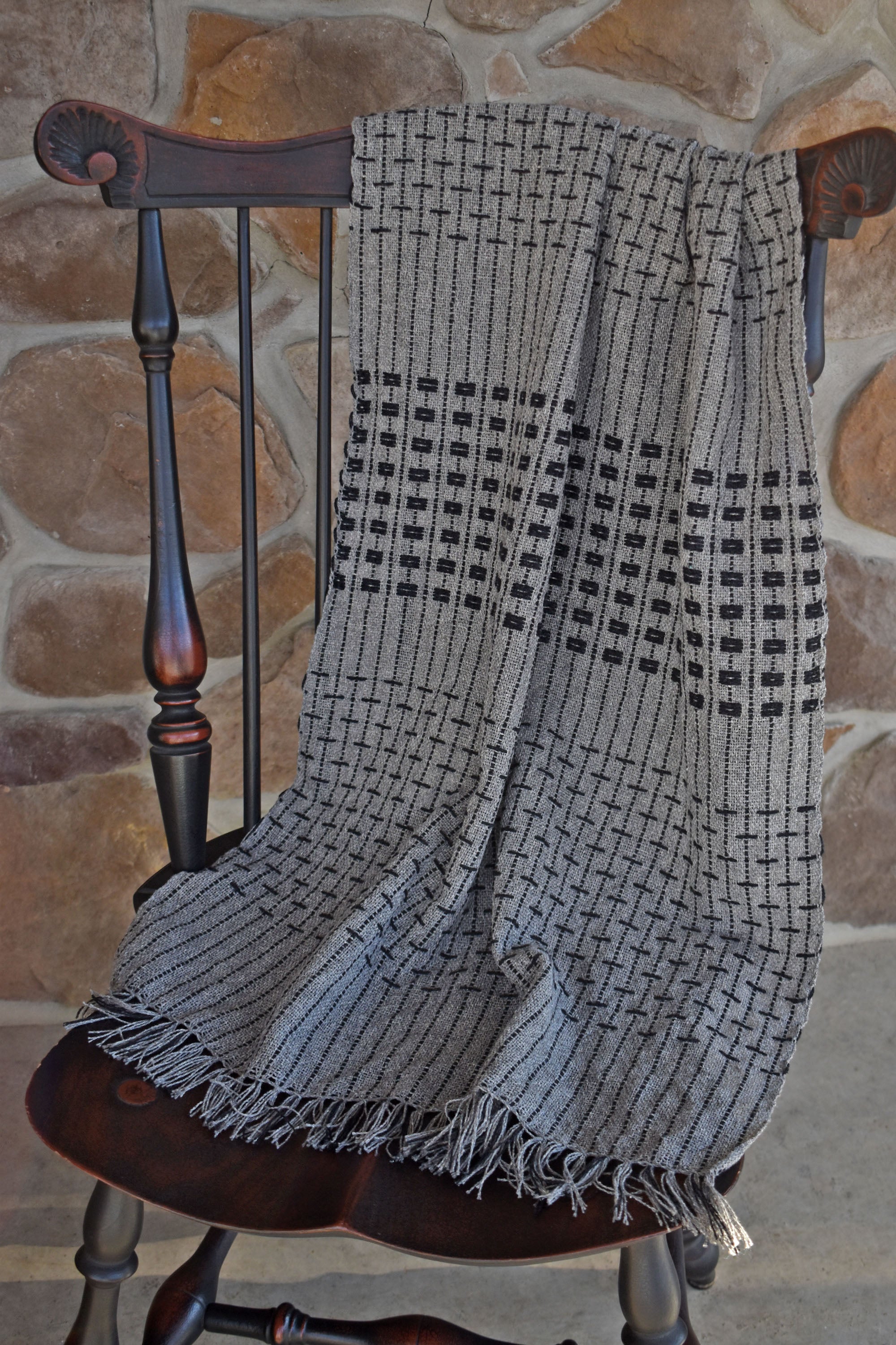 The women's handwoven gray shawl draped over a chain with a stone wall behind it. The eyelash fringe is also in view.