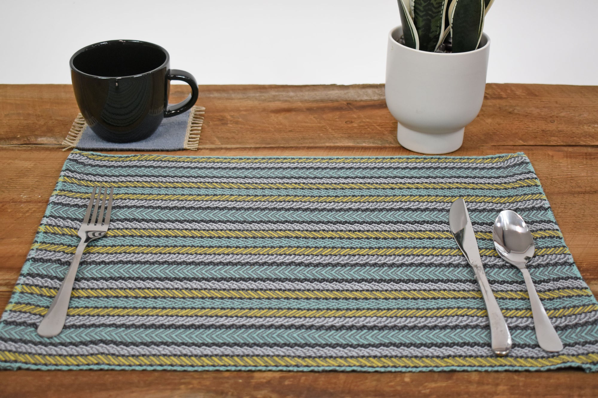 Striped placemat on a wooden table. Silverware is on top of the placemat. A mug and a plant is also in view.