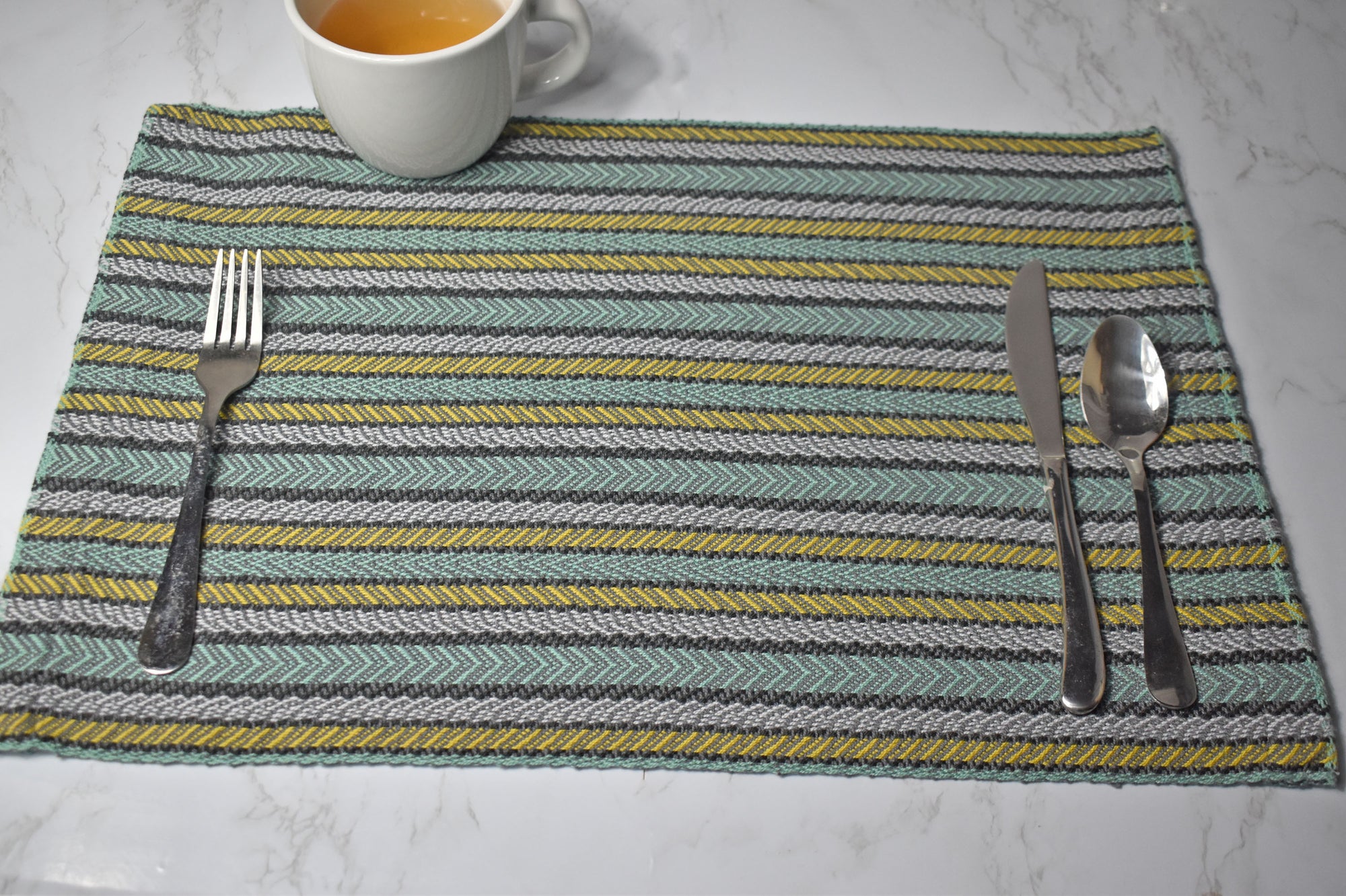 Striped placemat on a white table. There's silverware on top of the placemat. Also a cup of tea is in view.