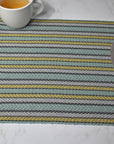 Striped placemat on a white table. There's silverware on top of the placemat. Also a cup of tea is in view.