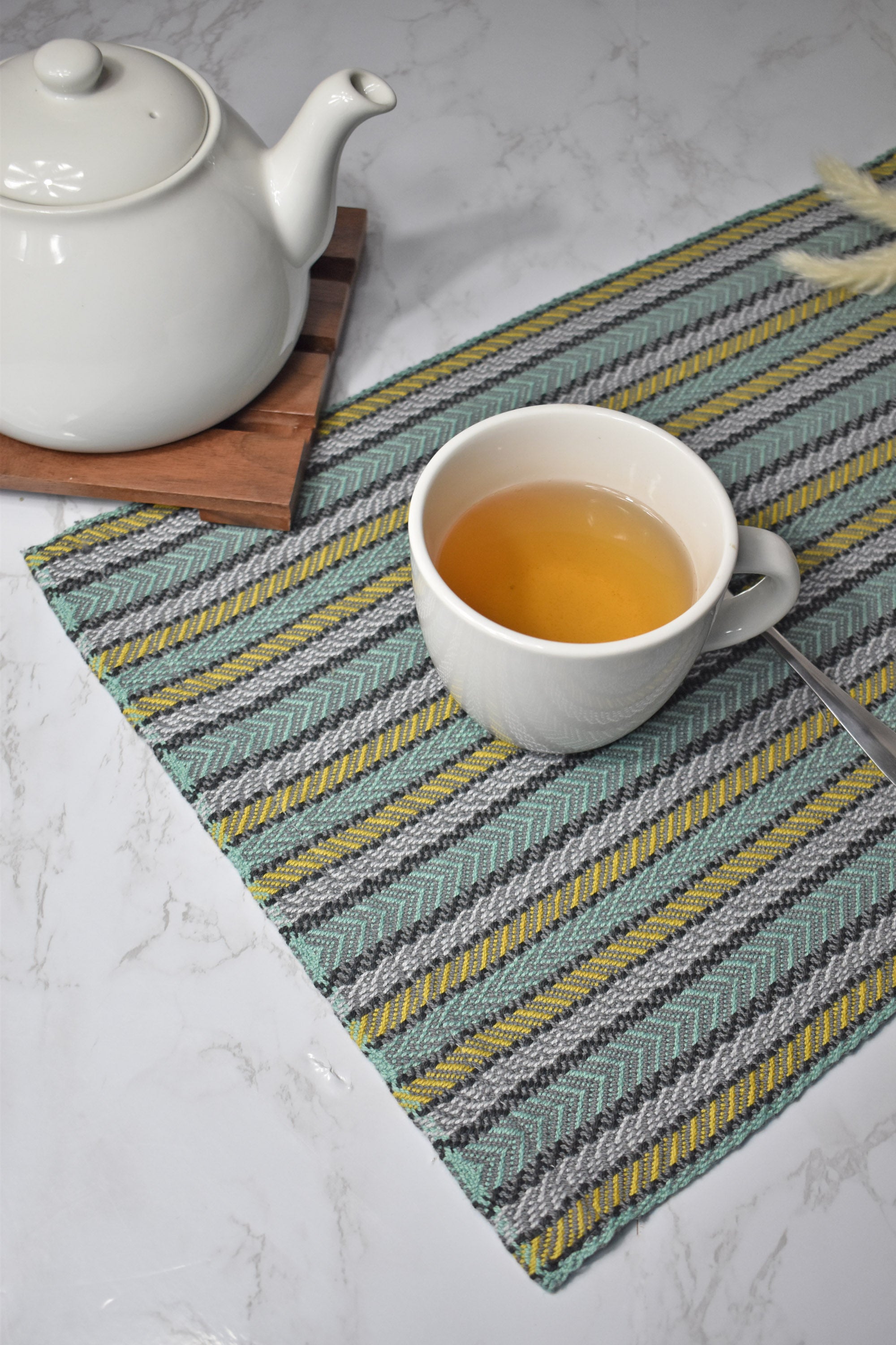 Green striped handwoven placemat on a white table. A cup of tea is on top of the placemat. A tea pot is in view too.