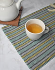 Green striped handwoven placemat on a white table. A cup of tea is on top of the placemat. A tea pot is in view too.