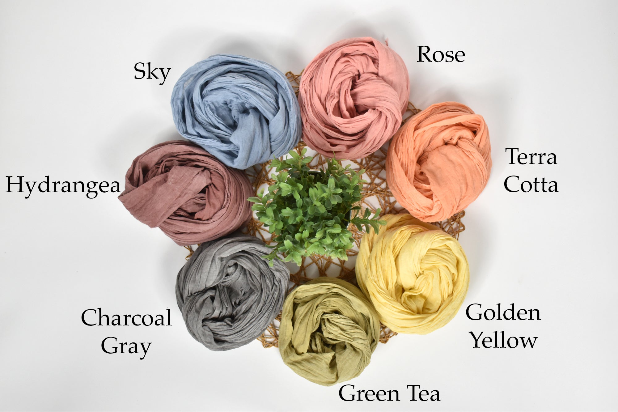 Naturally hand-painted scarves wrapped up laying on a table with a plant in the middle. Colors are labeled.