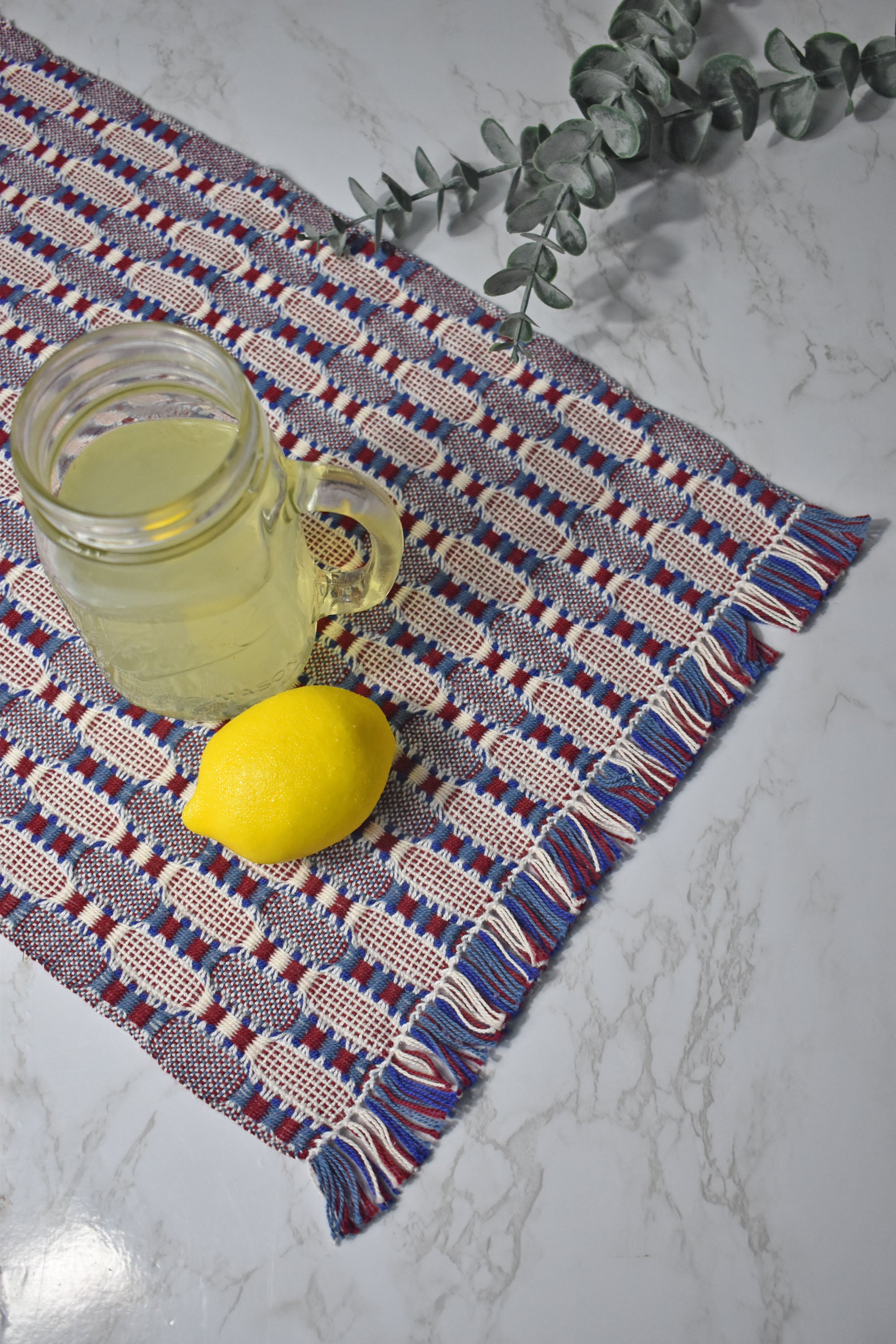 Patriotic handwoven organic cotton placemat on a white table. A lemon and a glass of lemonade on top of the placemat. Eucalyptus in view.