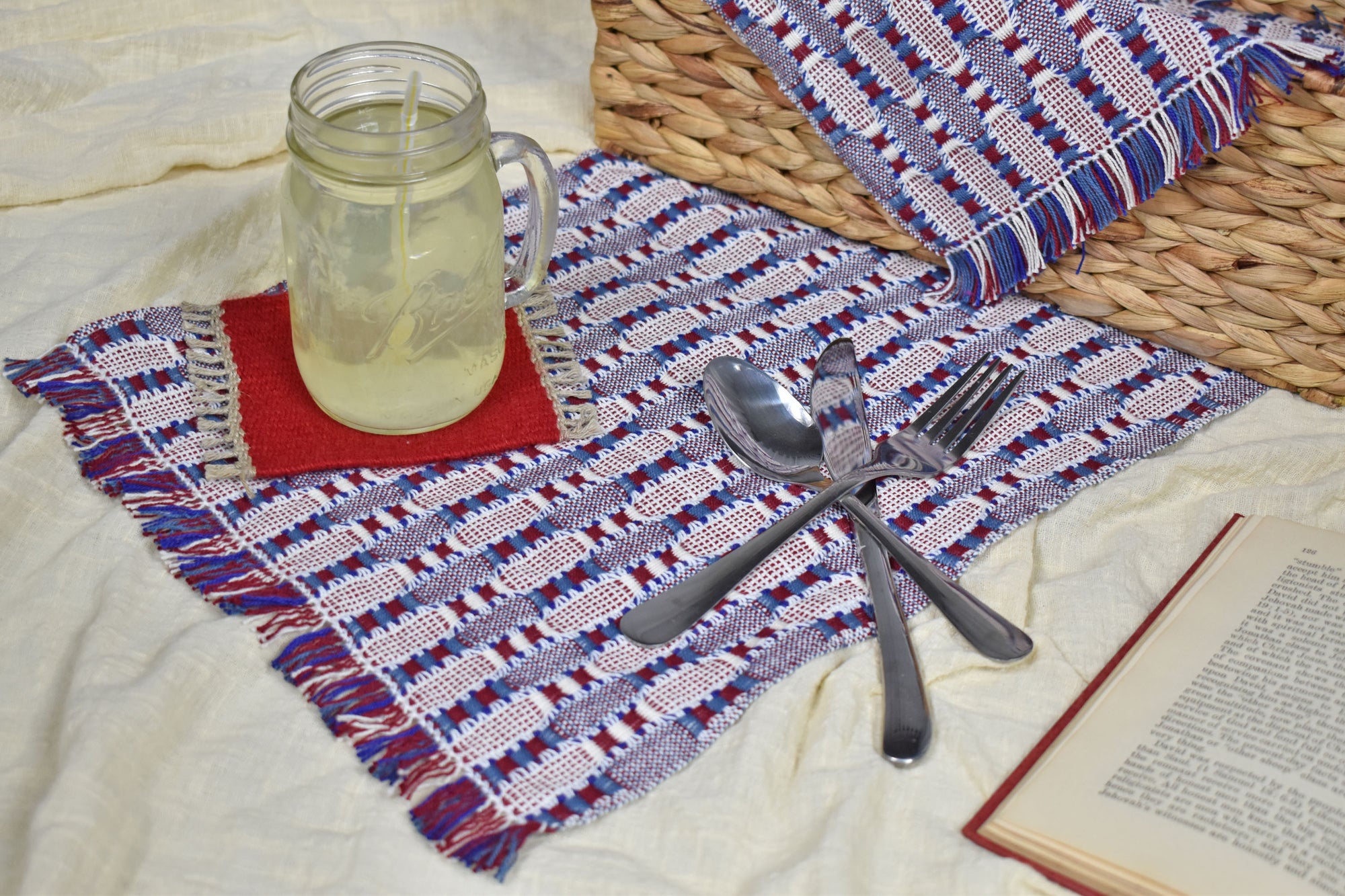 Patriotic placemat on top of a blanket with a picnic basket, glass of lemonade, silverware, and a book