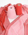 Pink + red scarf draped on a white table with the red and pink cones of yarns