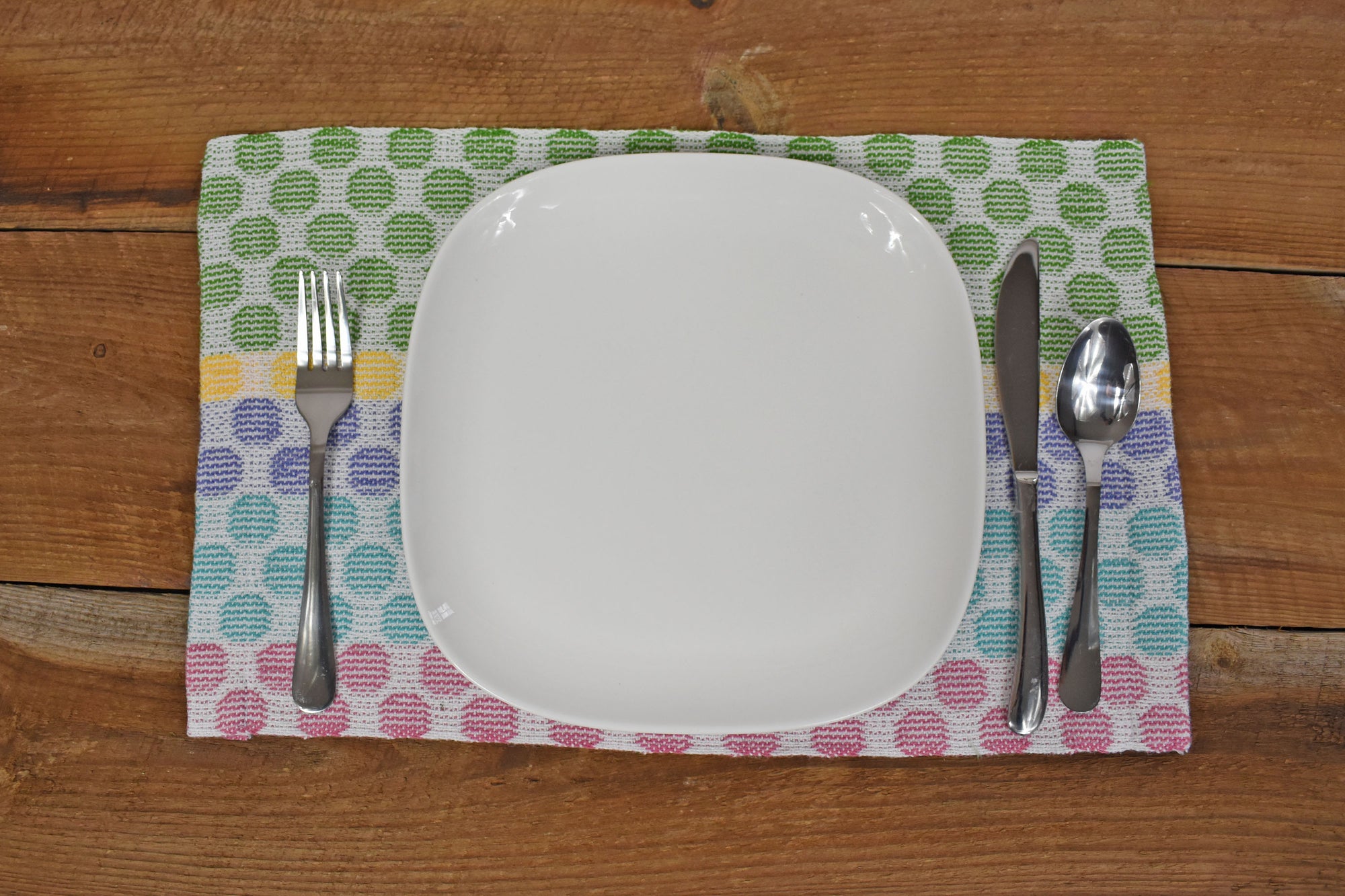 The polka dot placemat is laying on a wooden table. There&#39;s a white plate and silverware on top of the placemat.