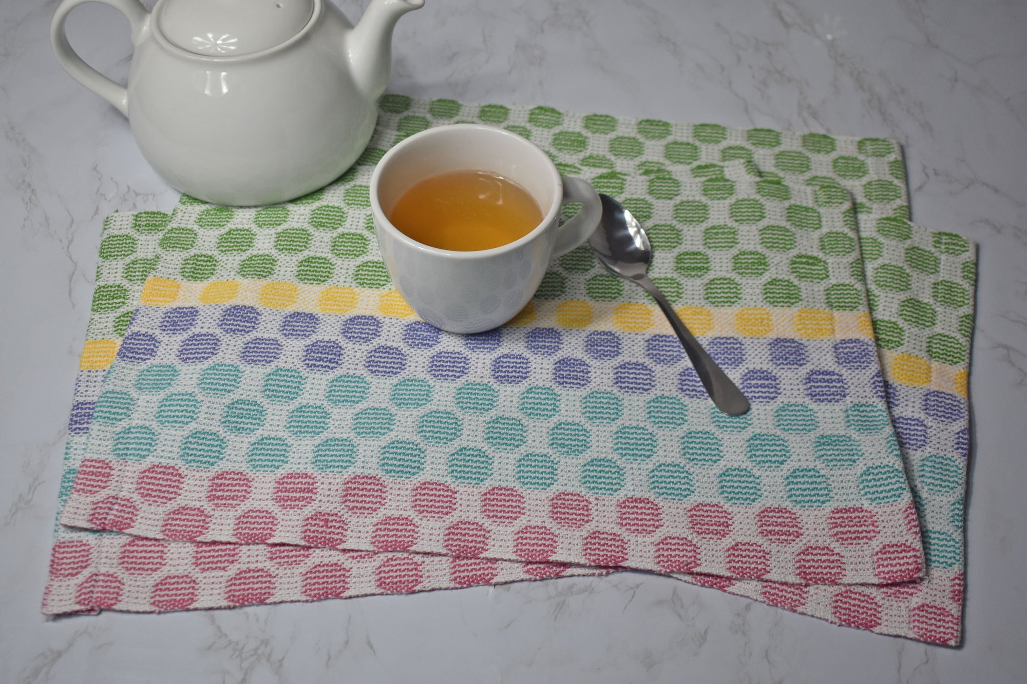 Several polka dot placemats on a white table. A cup of tea, spoon, and tea pot are also in view.