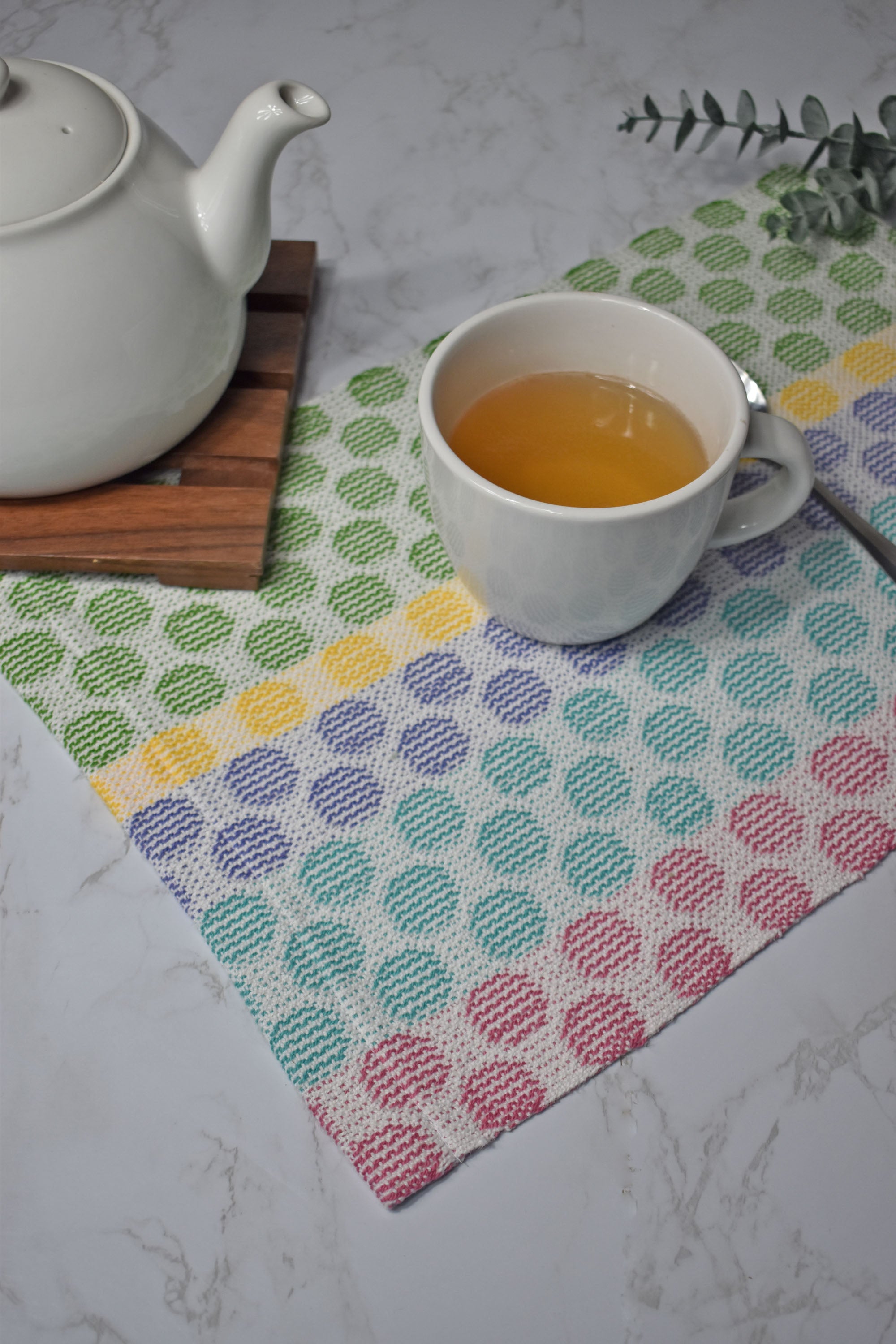 Polka dot handwoven placemat on a white table. There is a cup of tea and a tea pot on top of the placemat. Eucalyptus is in the background