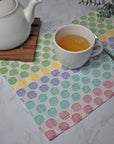 Polka dot handwoven placemat on a white table. There is a cup of tea and a tea pot on top of the placemat. Eucalyptus is in the background