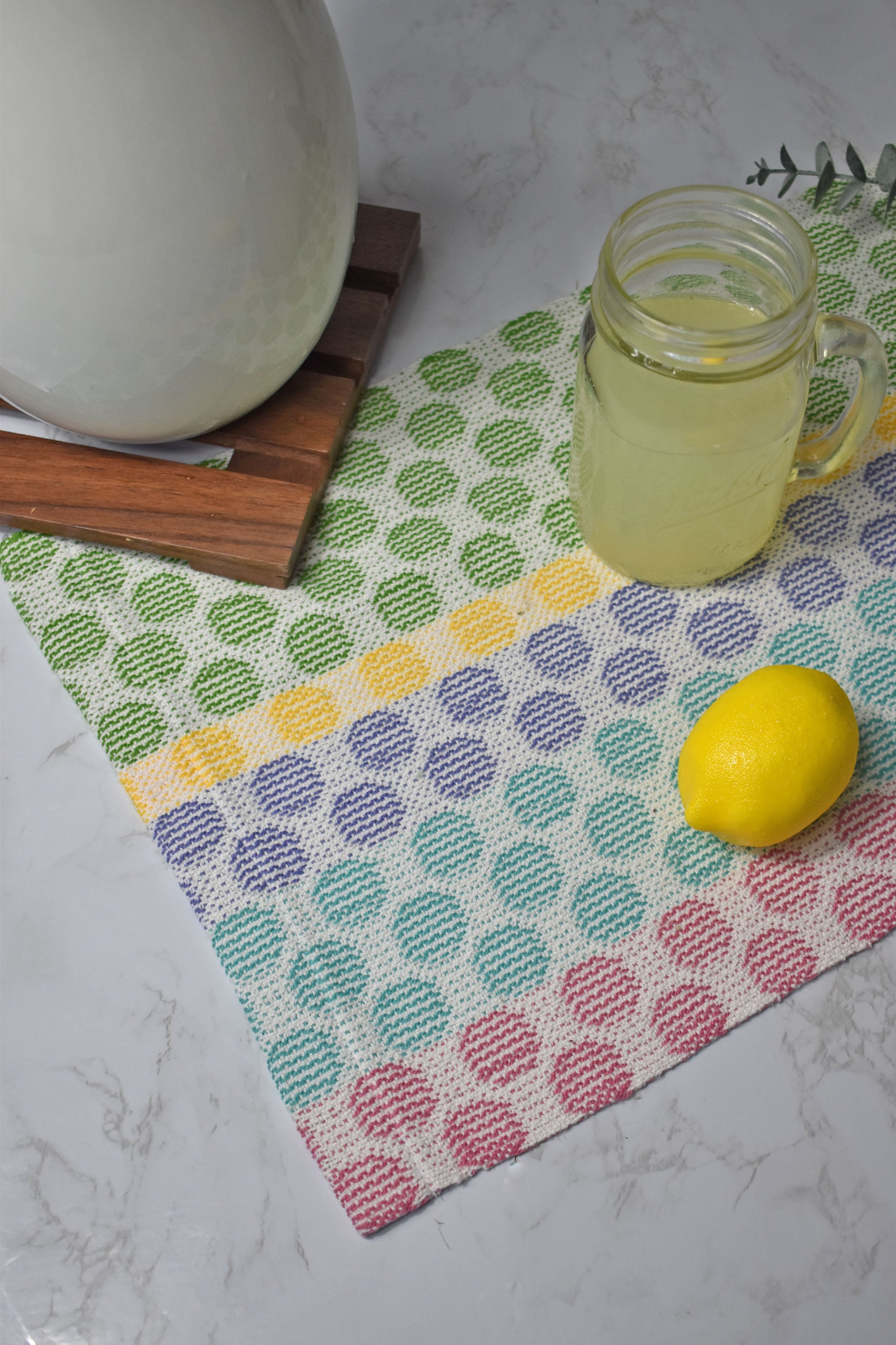 Handwoven placemat on a white table with a lemon, and a glass of lemonade on top of the placemat. A trivot and pitcher is in view.