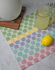 Handwoven placemat on a white table with a lemon, and a glass of lemonade on top of the placemat. A trivot and pitcher is in view.