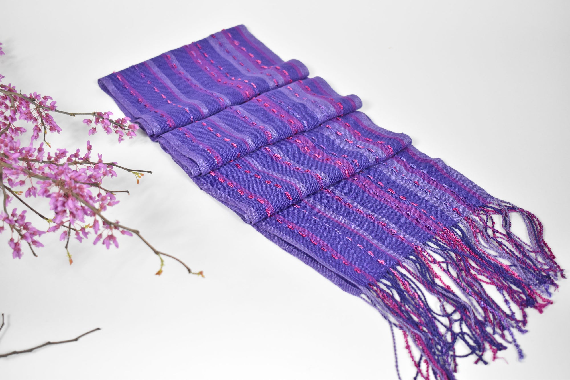 Handwoven purple scarf displayed on a white table with some purple flowers