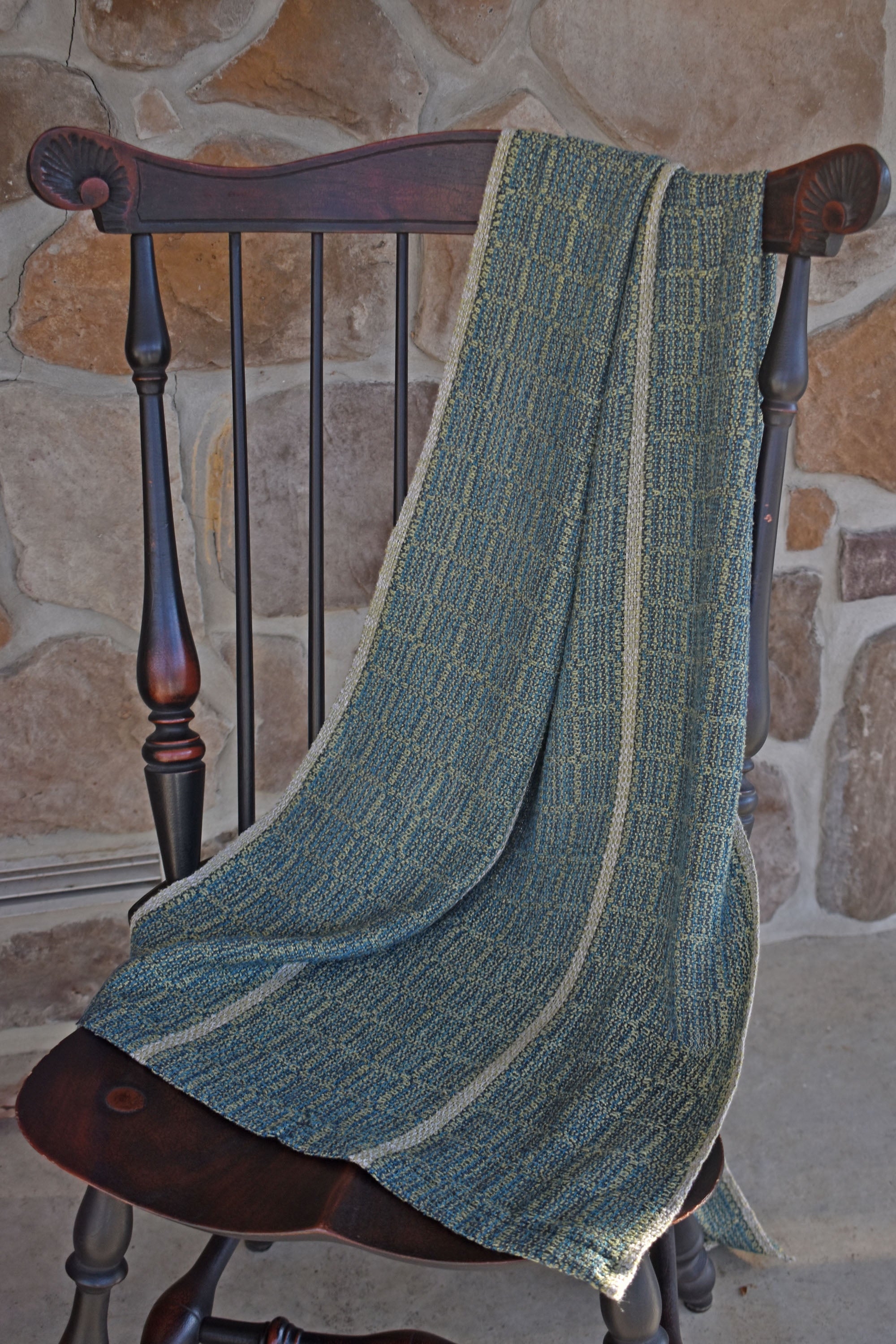 The handwoven blue and green scarf draped over a chair with a stone wall in the background.