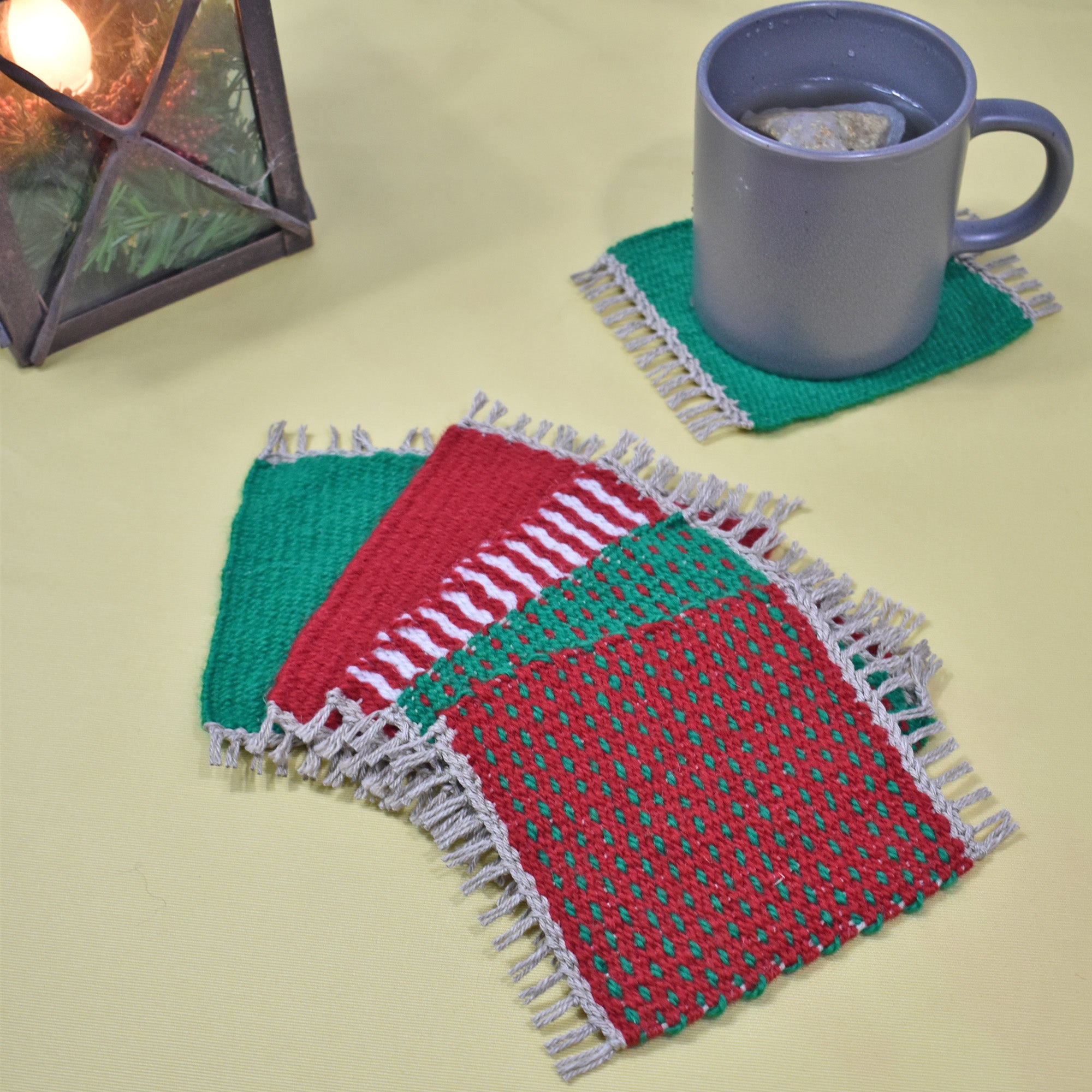 Handwoven Christmas mug rugs on a yellow table with a lantern and a cup of tea.