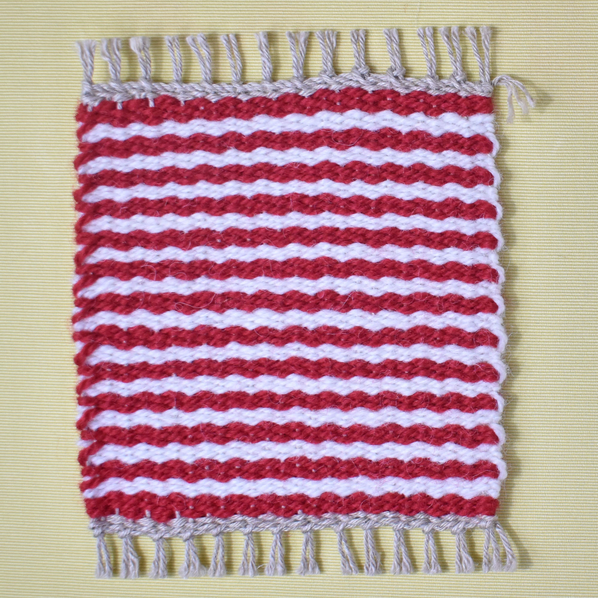 Red and white striped handwoven mug rug laying on a yellow table.